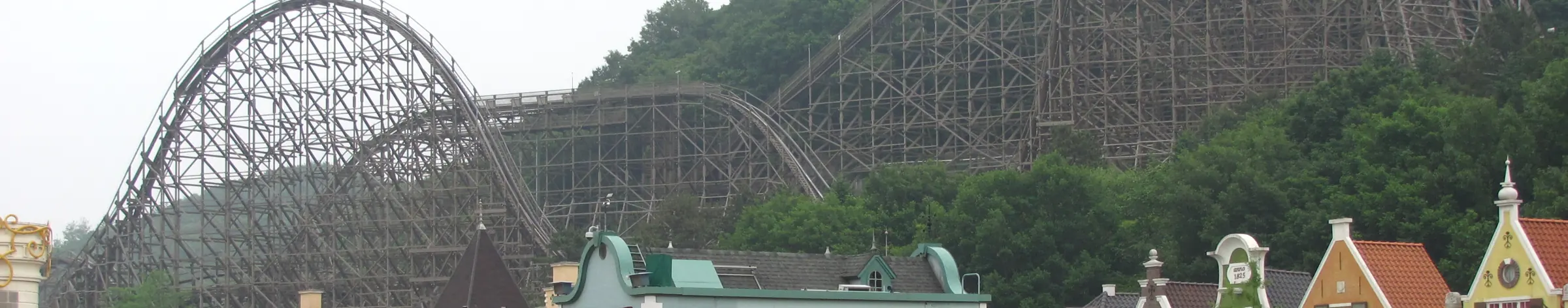 A wide view of a wooden roller coaster climbing a wooded hillside above a street of pastel European-style houses and flower beds; a general photograph of the coaster and its setting