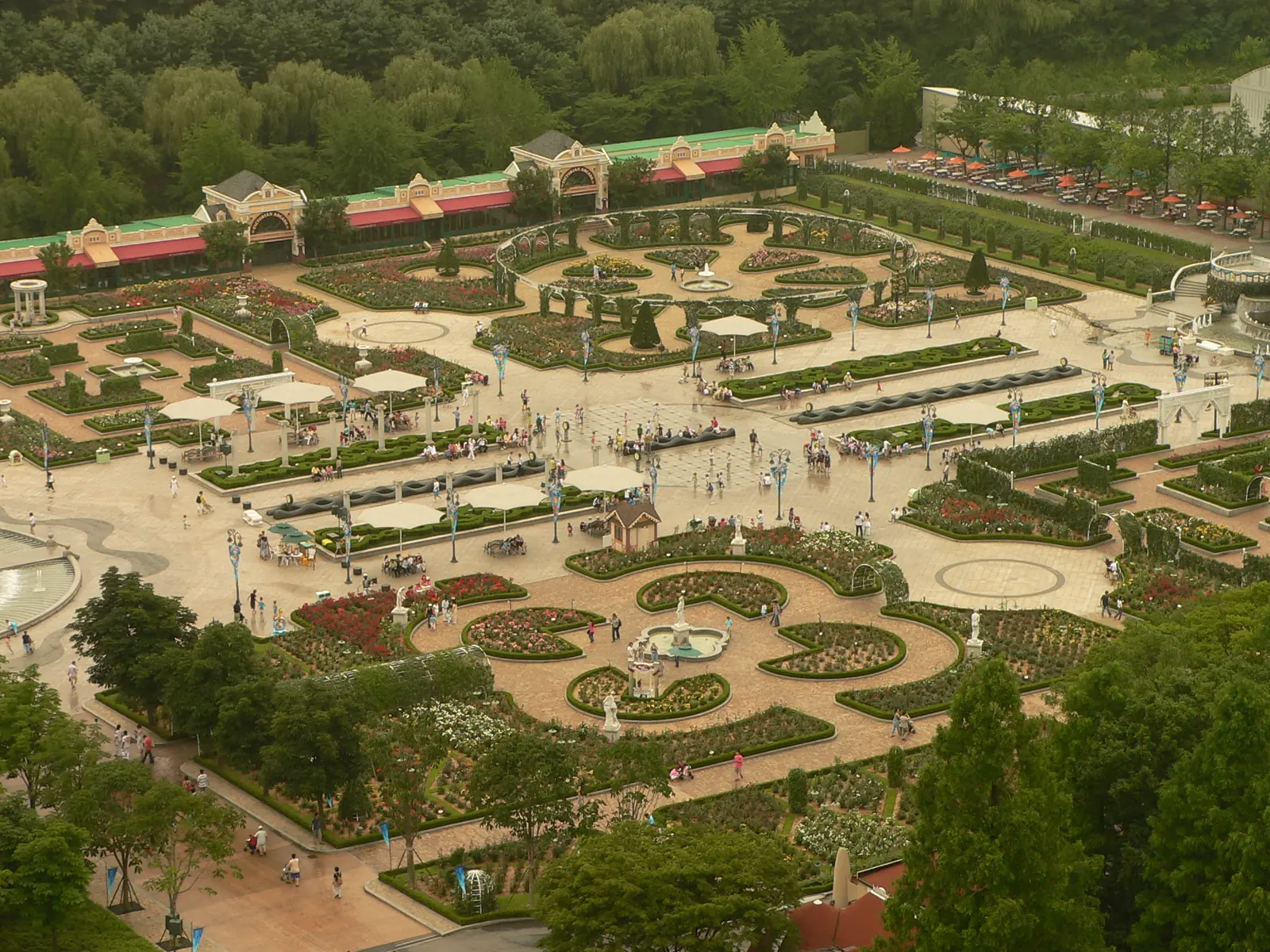 Formal flower gardens with curved paths, fountains and white umbrellas seen from above, with a long low building and trees behind