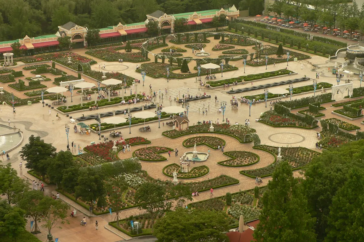 Formal flower gardens with curved paths, fountains and umbrellas seen from above, with a long low building and trees behind