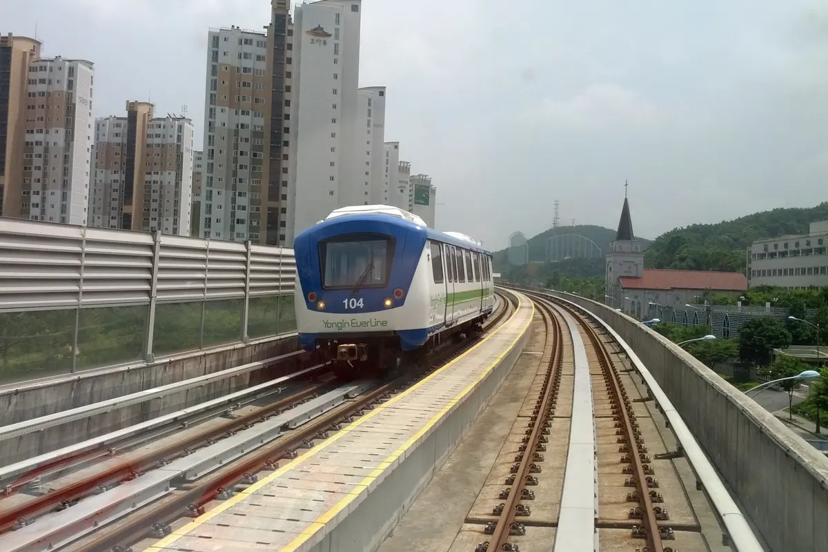 A white and blue Yongin Everline train with the number 104 on its front runs along an elevated concrete track beside residential towers, with wooded hills behind