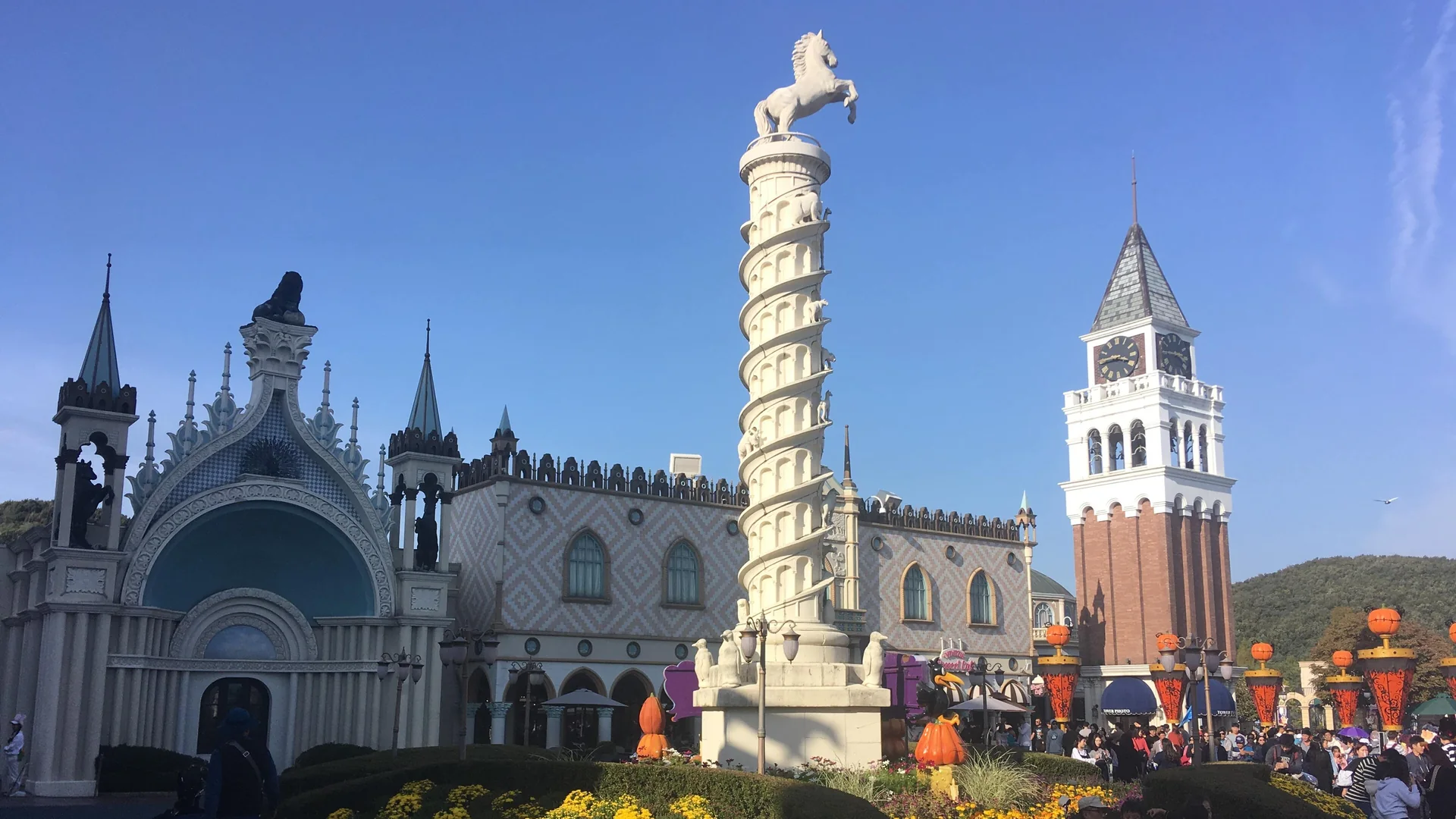 A tall white spiral column topped by a rearing horse stands in a plaza of pale Venetian-style buildings beside a red-brick clock tower under a deep blue sky, with orange pumpkin lanterns and yellow flowers at the base