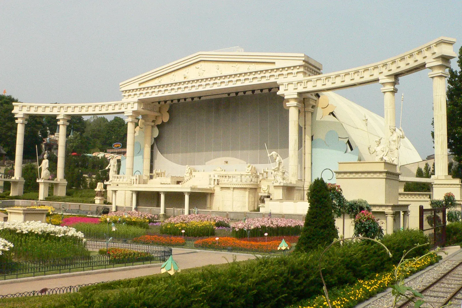 A white open-air stage with Greek-style columns and statues stands behind flower beds and a clipped hedge under a hazy sky